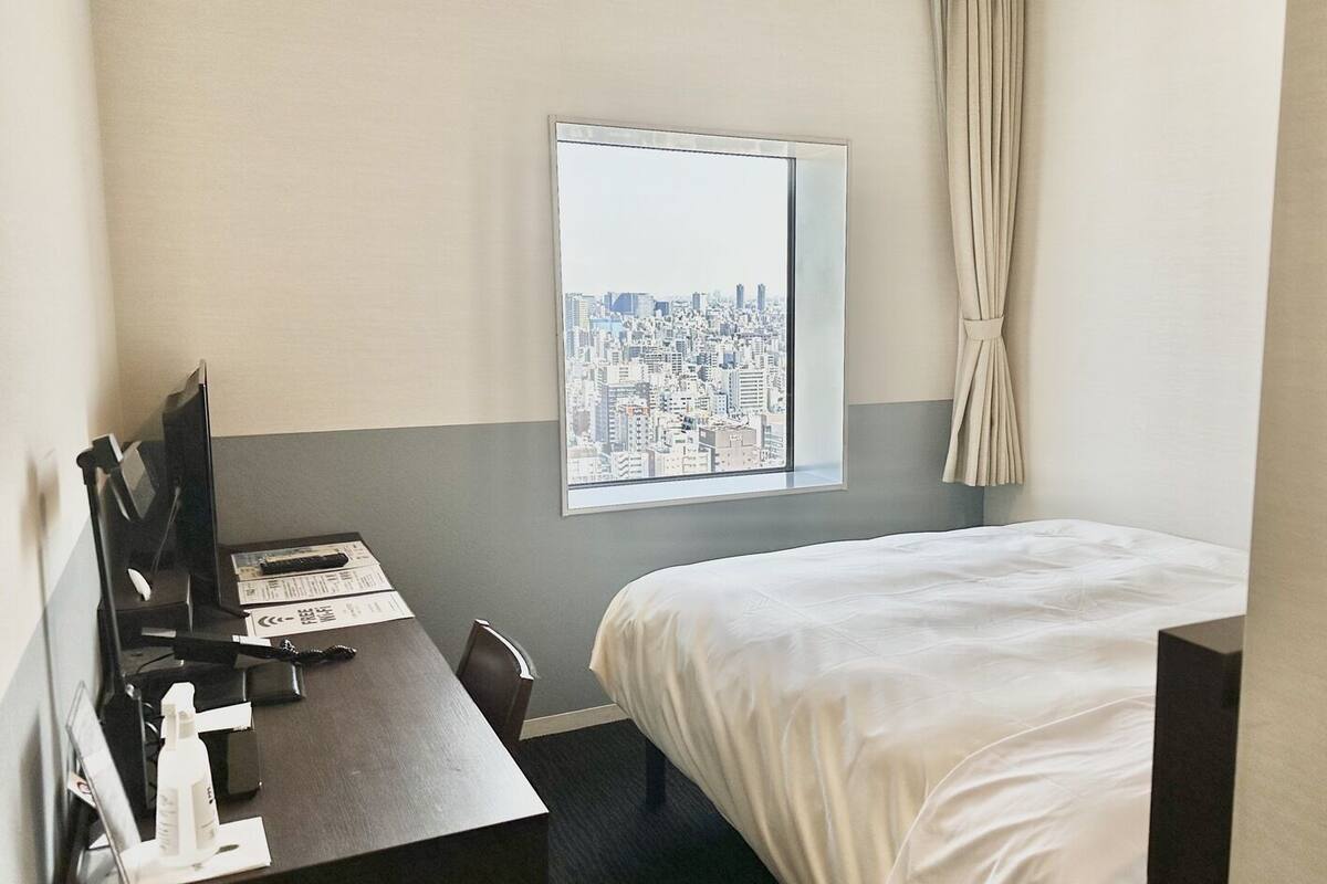 Een hotelkamer met een bed, een bureau met een computer, een raam met uitzicht op de stad en een gordijn.