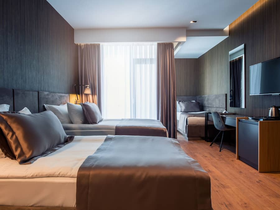 Habitación de hotel moderna con dos camas, un escritorio, una silla, un televisor y un ventanal grande con cortinas.