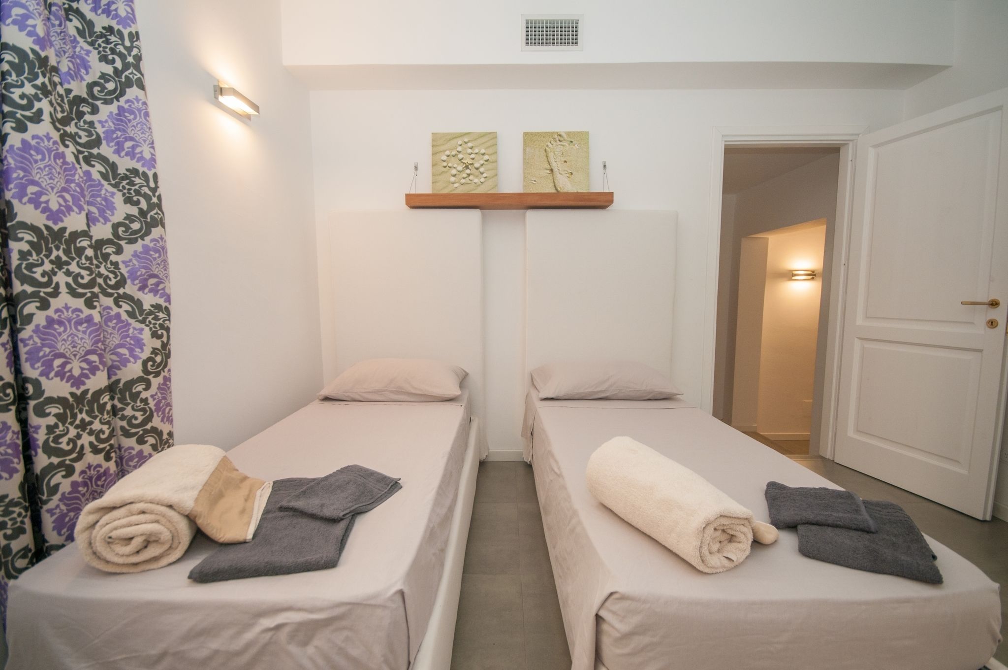 A hotel room with two single beds, a small shelf with artwork, a door, and a patterned curtain.