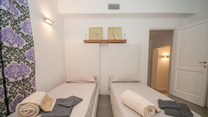 A hotel room with two single beds, a small shelf with artwork, a door, and a patterned curtain. - Oleandri 101 (Arzachena)