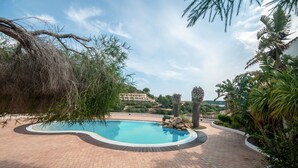 A swimming pool with a brick pathway, surrounded by greenery and a building in the background. - Oleandri 238 (Arzachena)