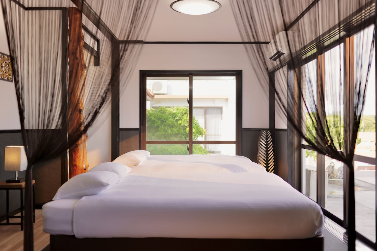 Habitación de hotel moderna con una cama grande, una mesita de noche, una lámpara y vistas al verdor a través de un amplio ventanal.