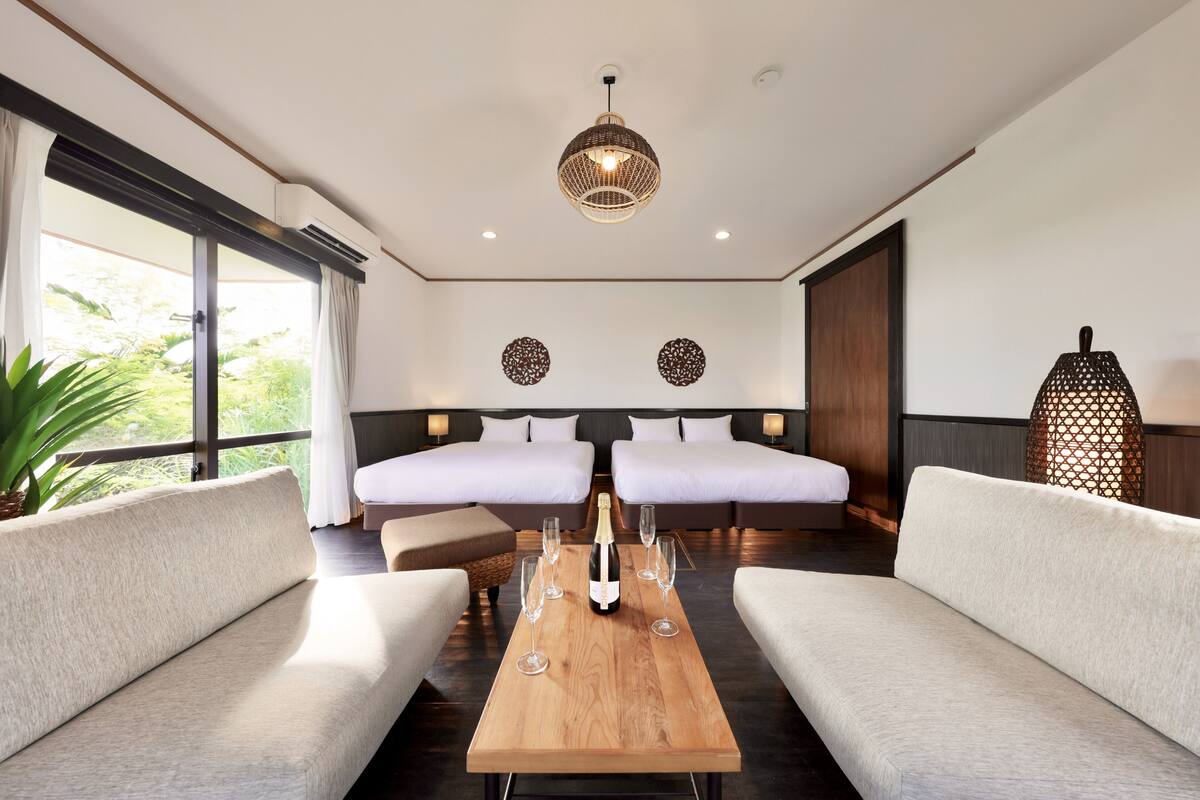 Une chambre d’hôtel moderne avec deux lits, une table en bois avec des verres et une bouteille, et une grande fenêtre avec des rideaux.
