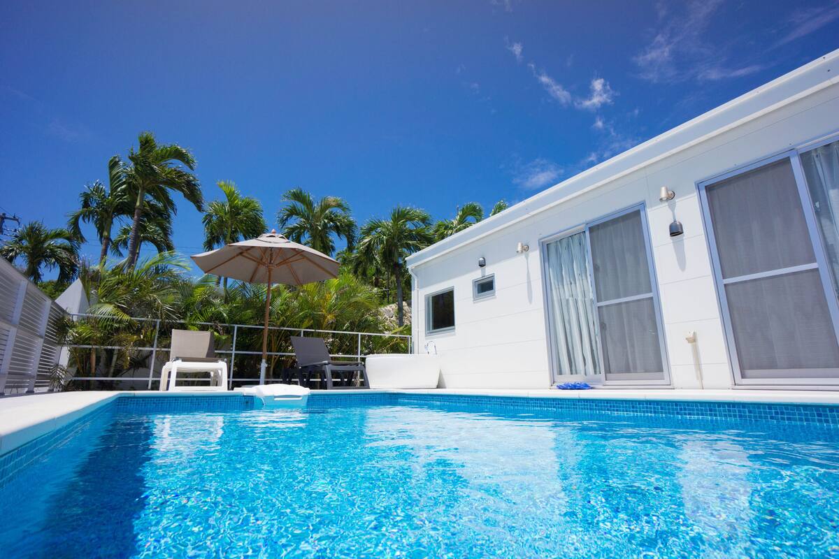 Une piscine à l’eau d’un bleu cristallin, entourée d’un bâtiment blanc doté de grandes fenêtres et d’un espace abrité avec des chaises et un parasol.
