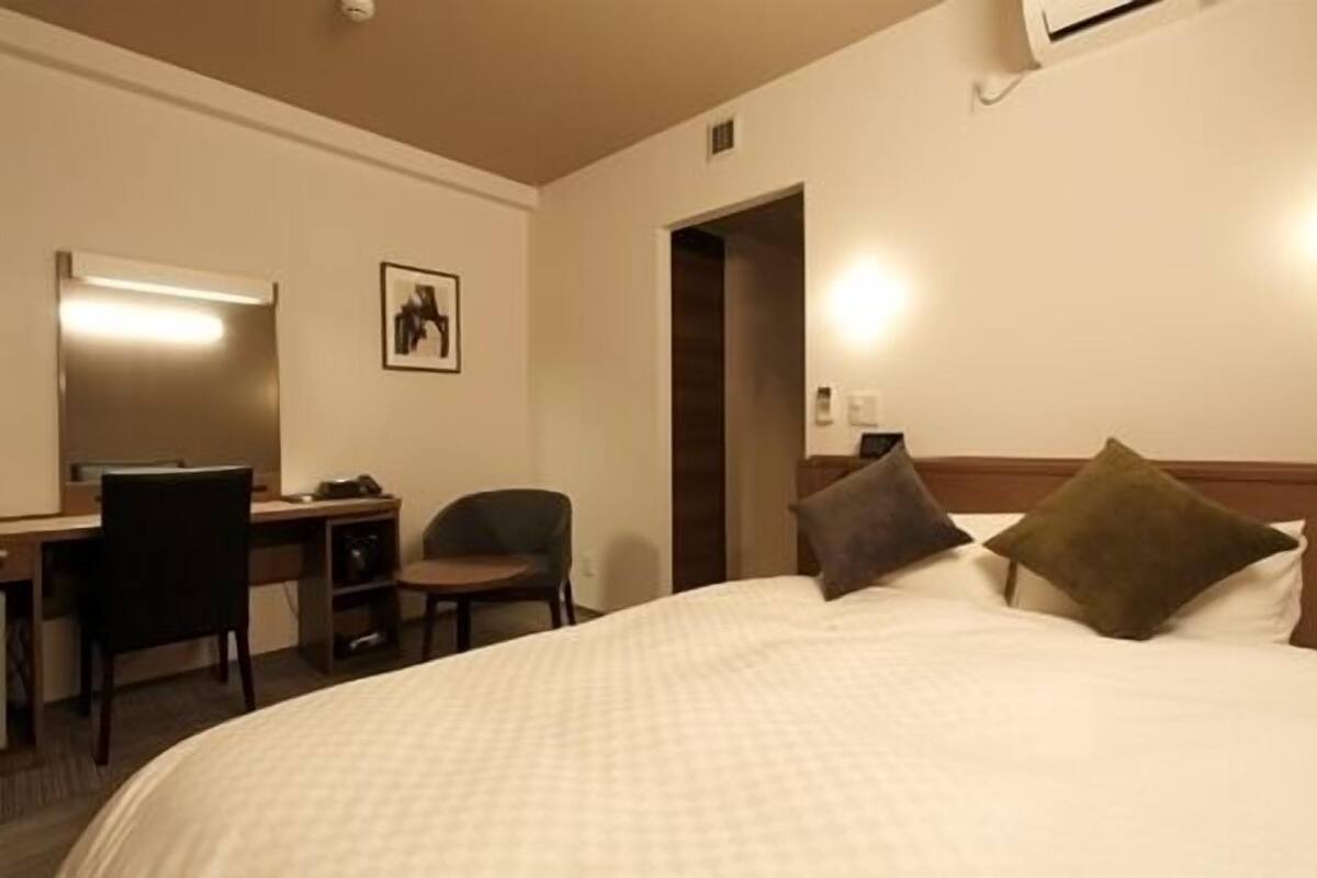 A hotel room with a bed, a desk with a chair, a picture on the wall, and an air conditioning unit.
