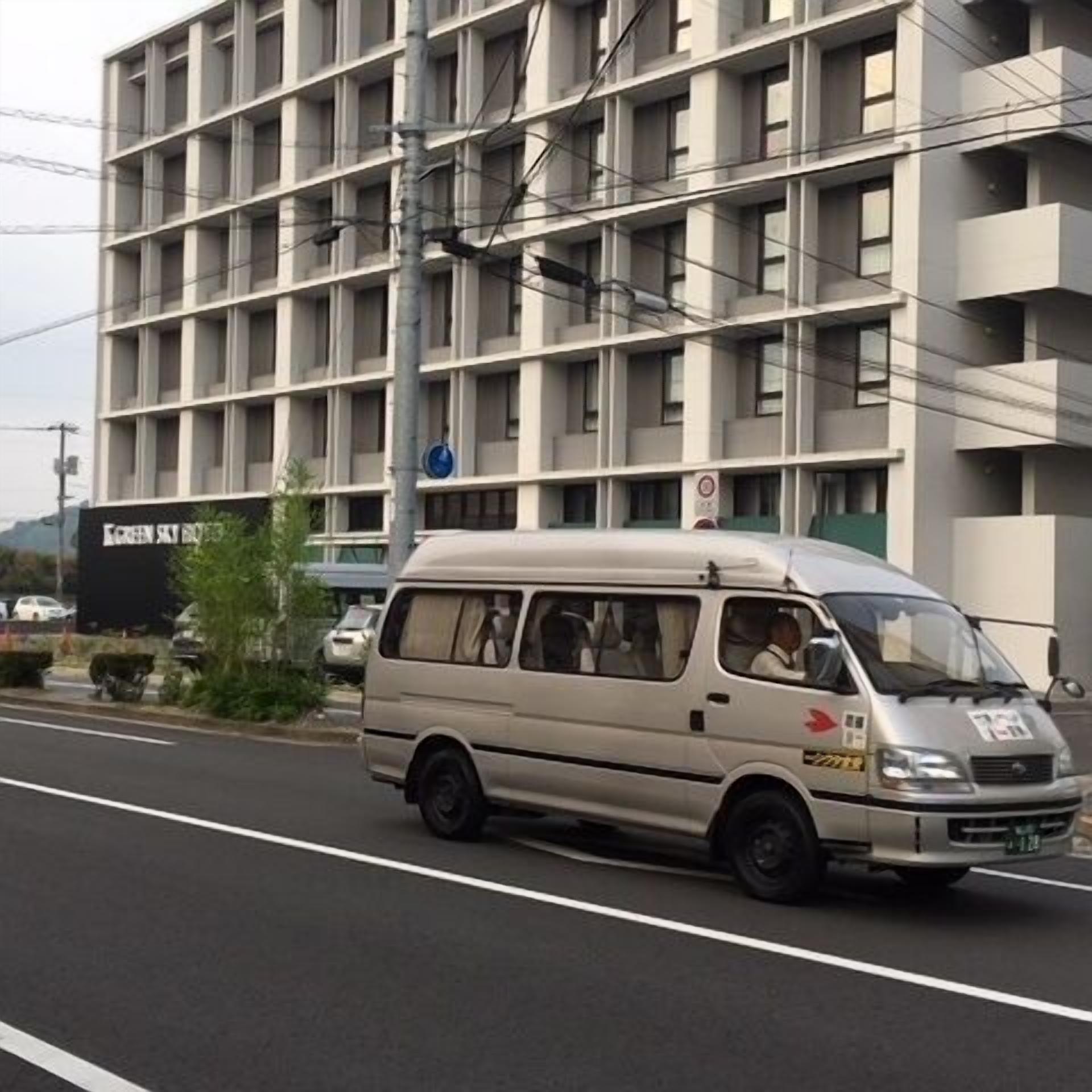 Um edifício com vários andares e fachada uniforme, uma carrinha estacionada na rua e uma placa com texto parcialmente visível.