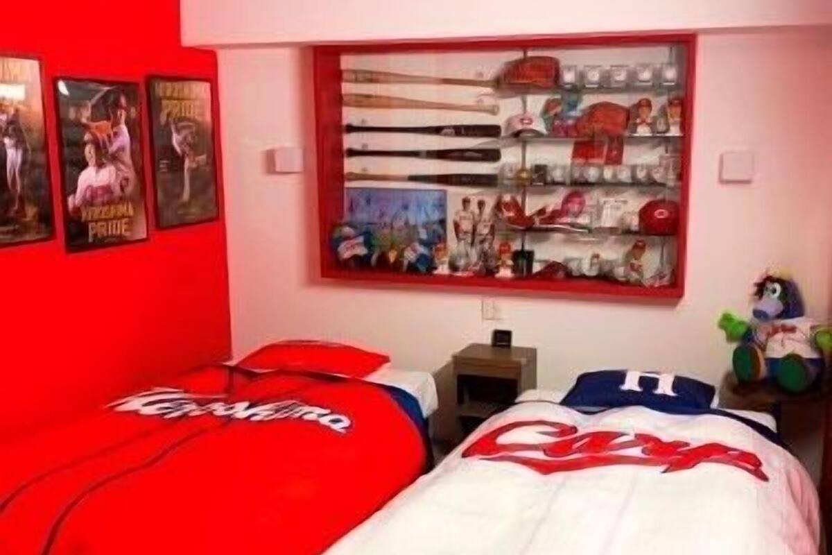 A bedroom with two beds, a window with a view of a cityscape, and a shelf with various items.