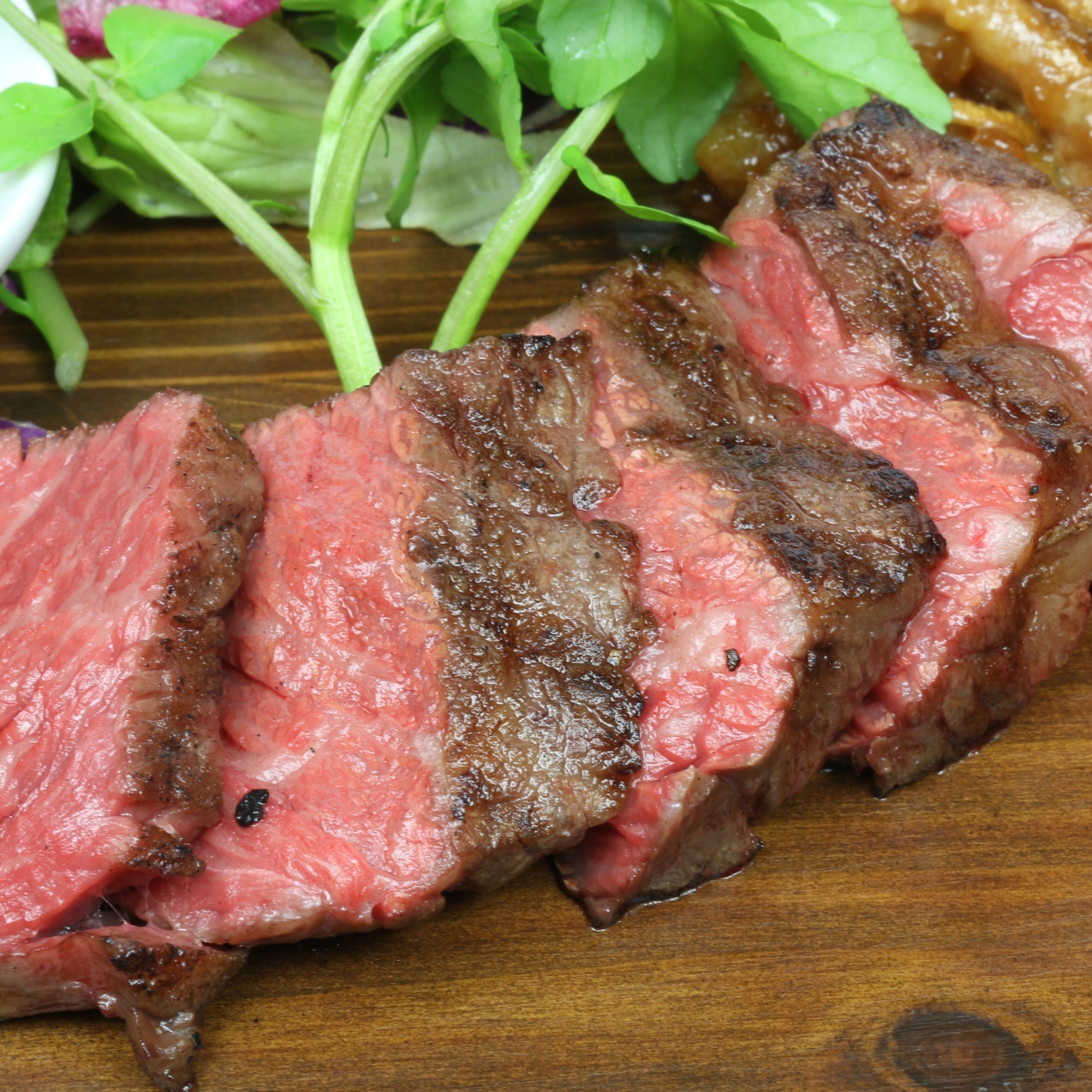 Un bistec con centro rosado y exterior quemado, servido sobre una tabla de madera con vegetales de hoja verde.