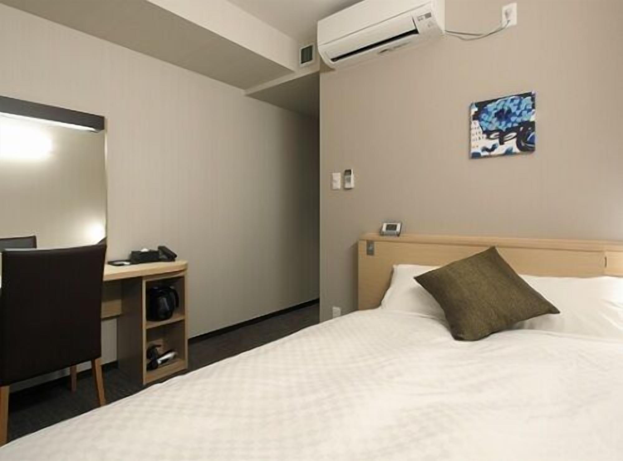 Een hotelkamer met een bed, een bureau met een stoel, een airconditioningapparaat en een aan de muur bevestigd telefoontoestel.