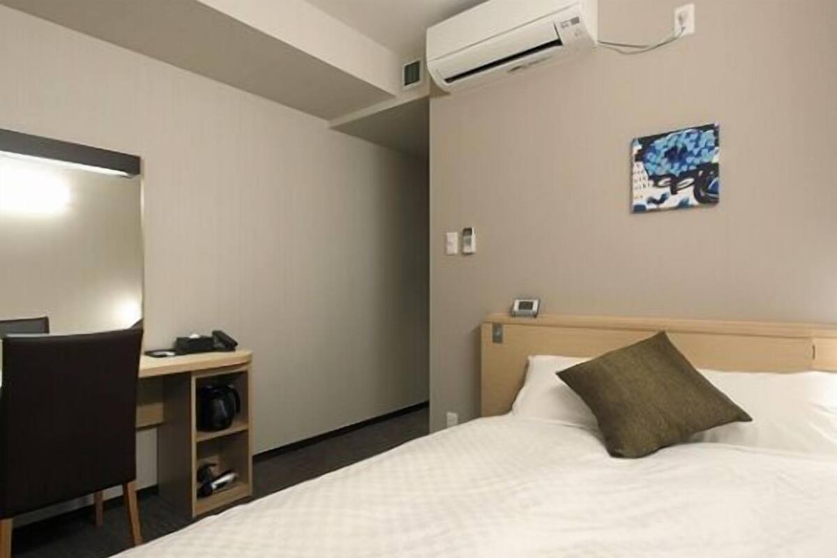 A hotel room with a bed, a desk with a chair, an air conditioning unit, and a wall-mounted phone.