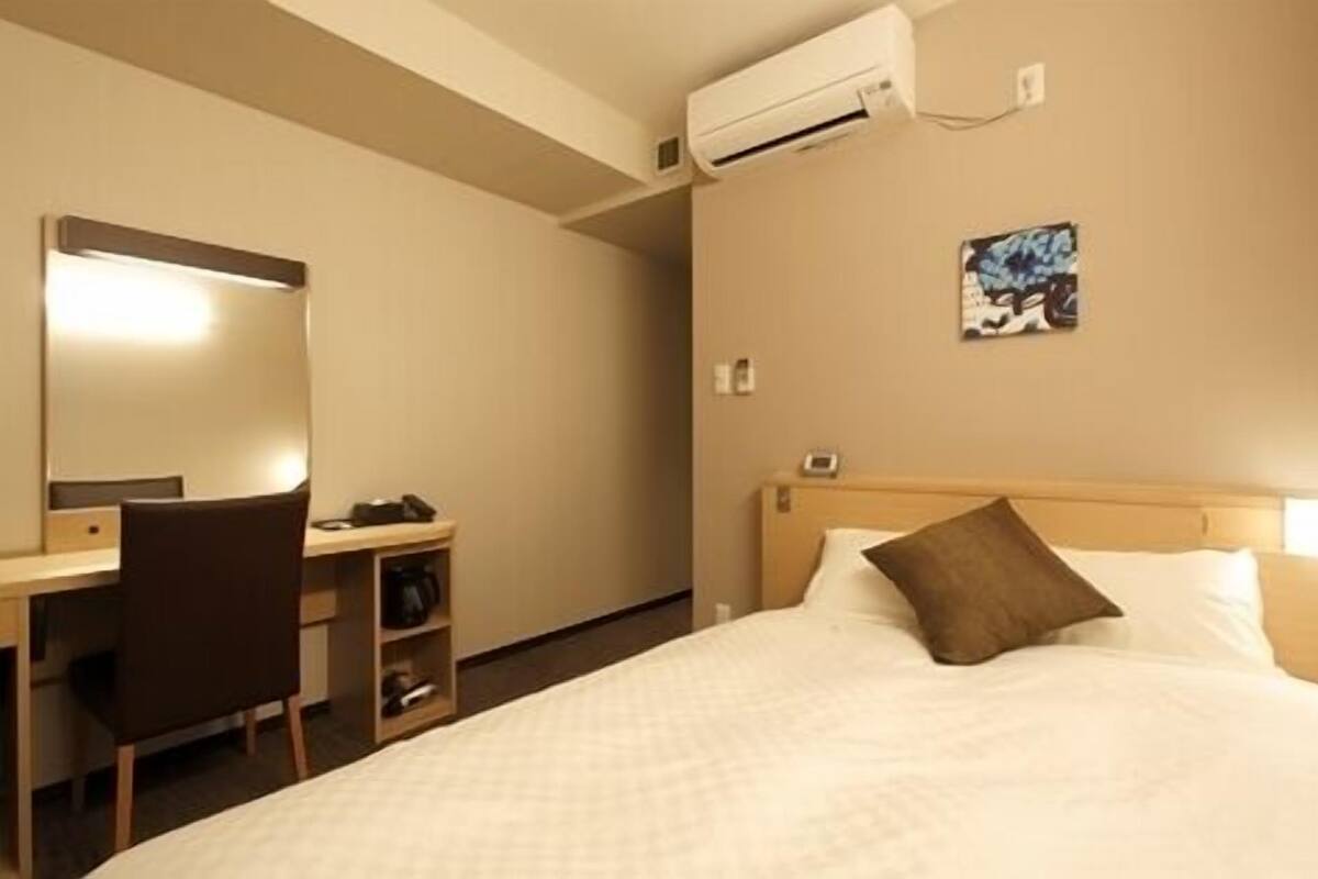 A hotel room with a bed, a desk with a chair, an air conditioning unit, a painting on the wall, and a nightstand.