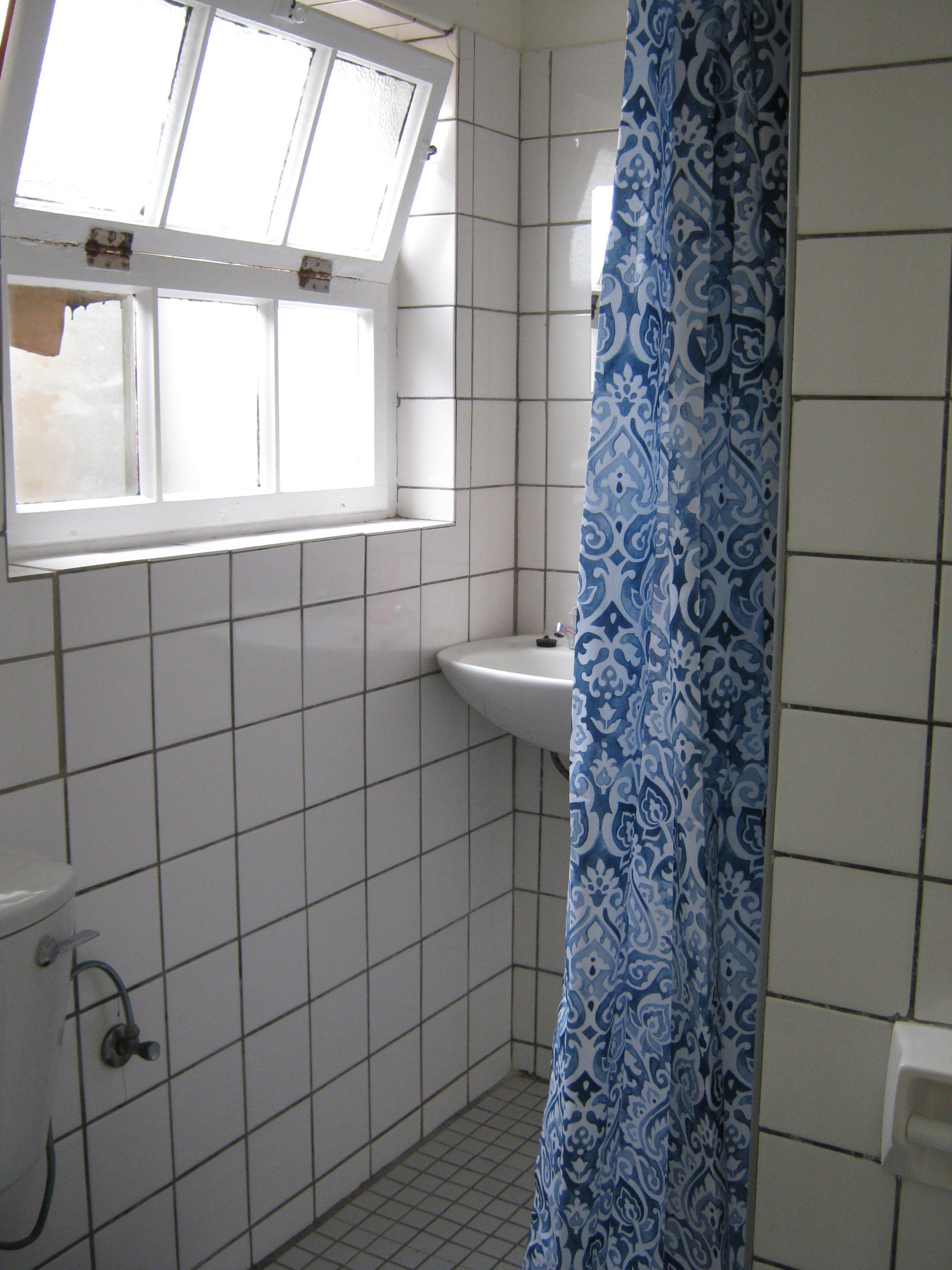 A small bathroom with white tiles, a window, a sink, and a shower curtain.