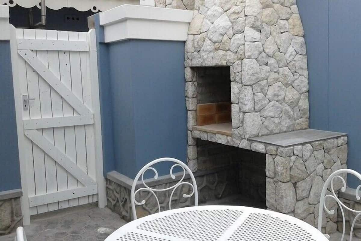 A stone-built outdoor fireplace with a tiled hearth, surrounded by a blue wall and a white gate.