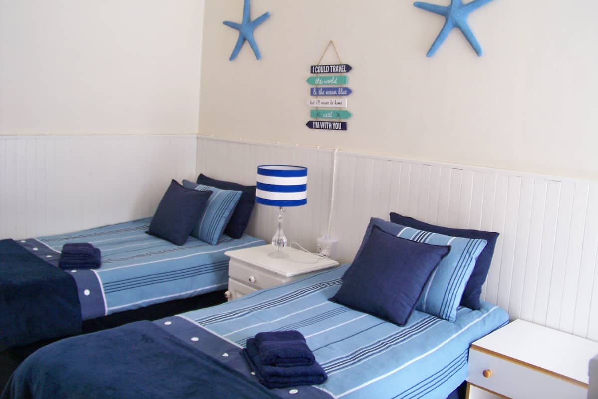 A room with two beds, each with matching blue bedding and pillows. There is a bedside table with a lamp and a small shelf. The walls are painted white, and there are starfish decorations on the wall.