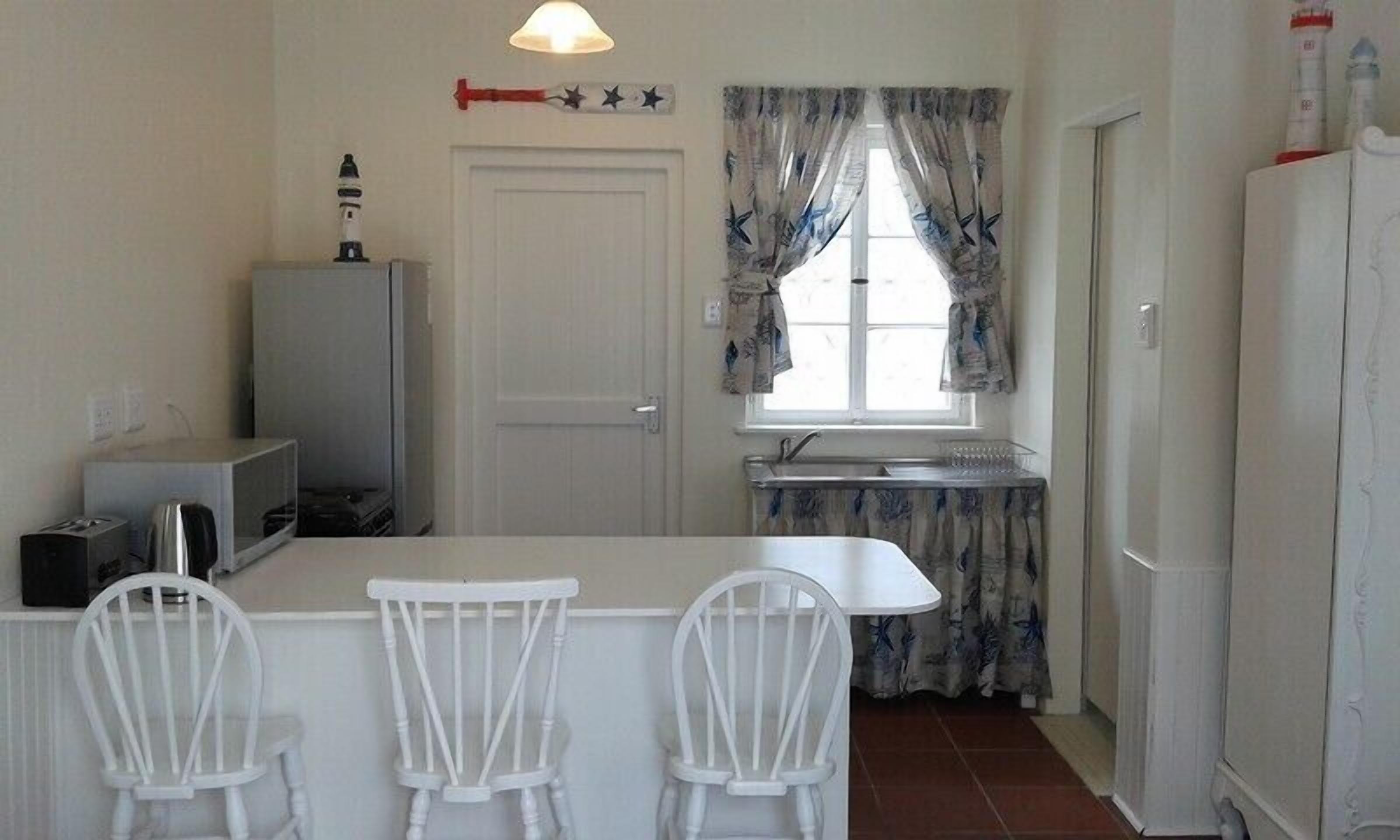 Una pequeña cocina con un mostrador blanco, tres sillas blancas, un refrigerador, un microondas, un lavamanos y una ventana con cortinas.