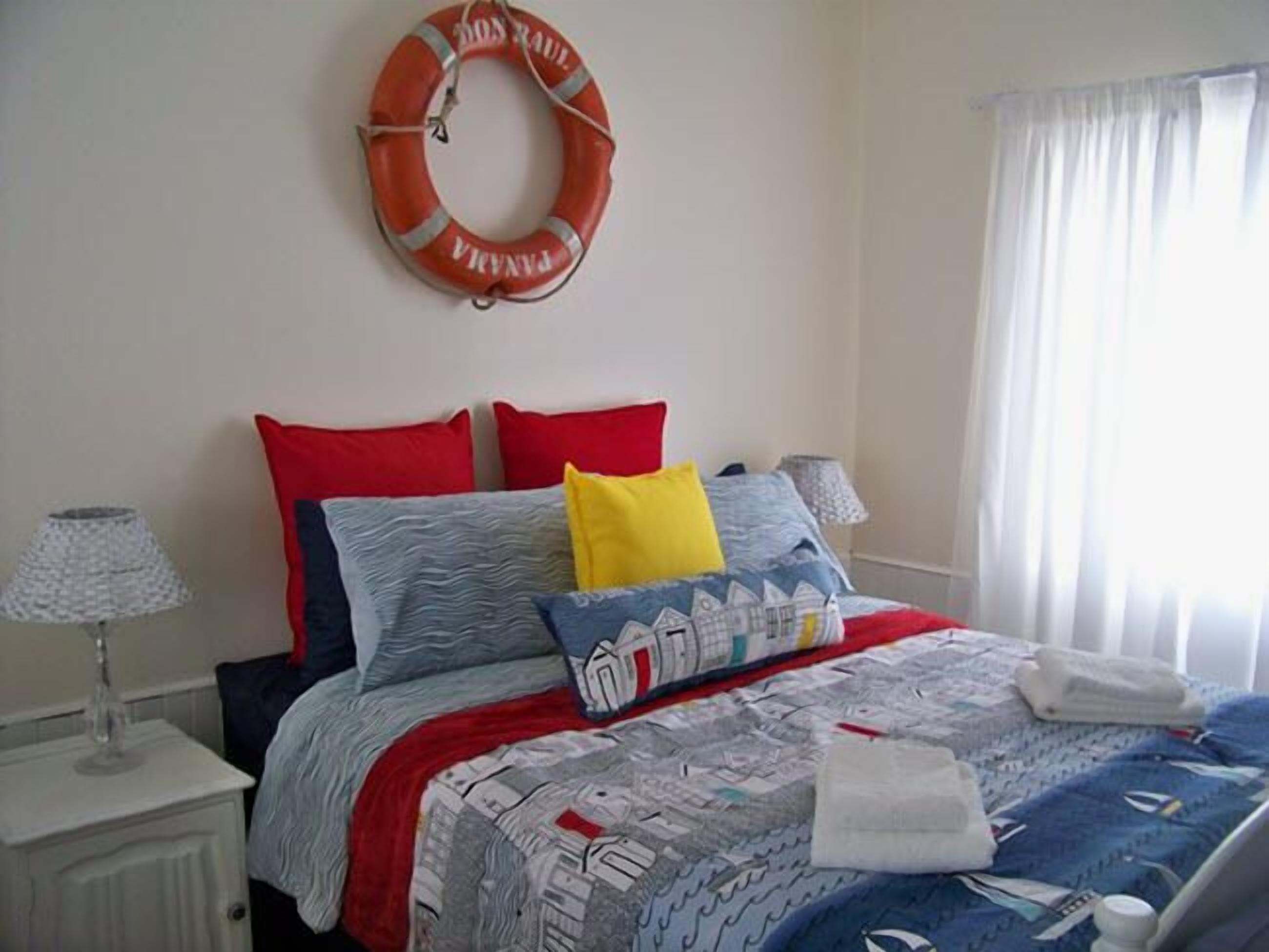 Un lit bien fait, recouvert d’une couvre-lit à motifs, avec deux oreillers rouges et un oreiller jaune. Un gilet de sauvetage est accroché au mur, au-dessus du lit.