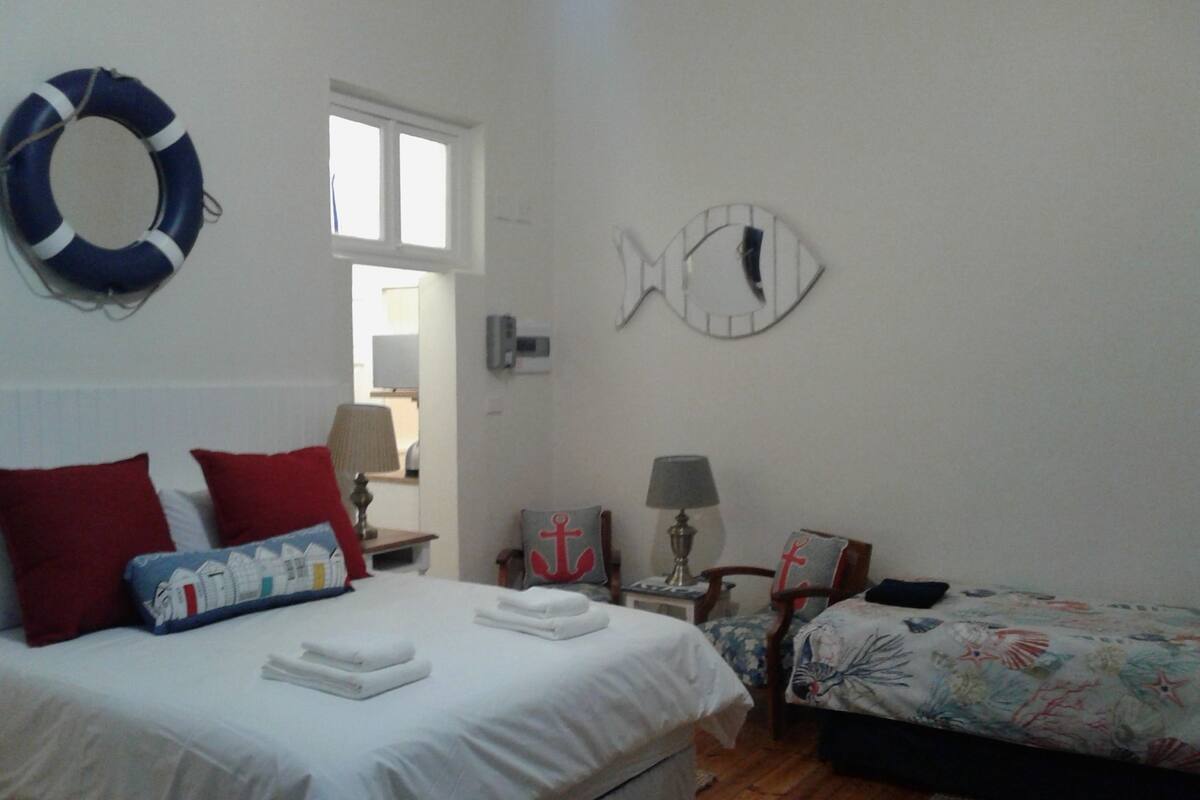 A bedroom with a bed, two bedside tables, a nightstand, a fish-shaped wall decoration, a life ring, and a window.