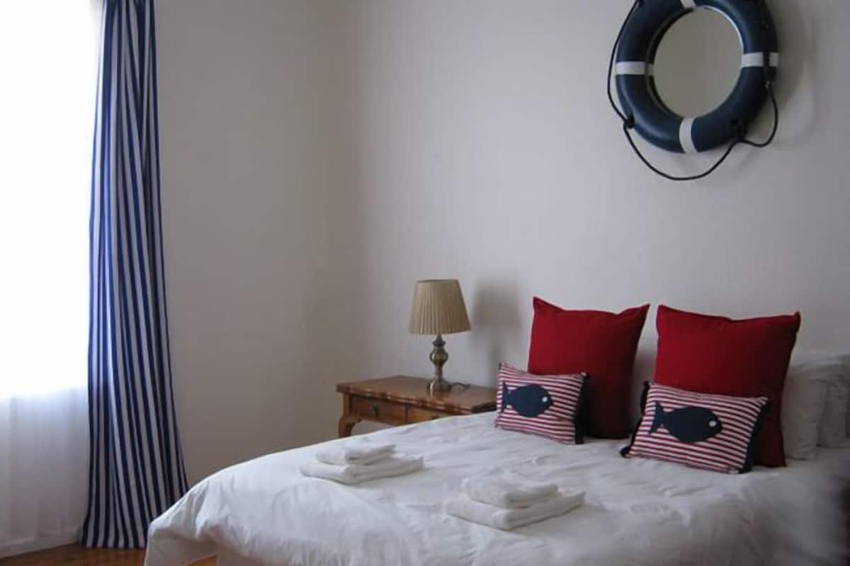 A neatly made bed with white linens and two red pillows, a bedside lamp, and a life ring on the wall.