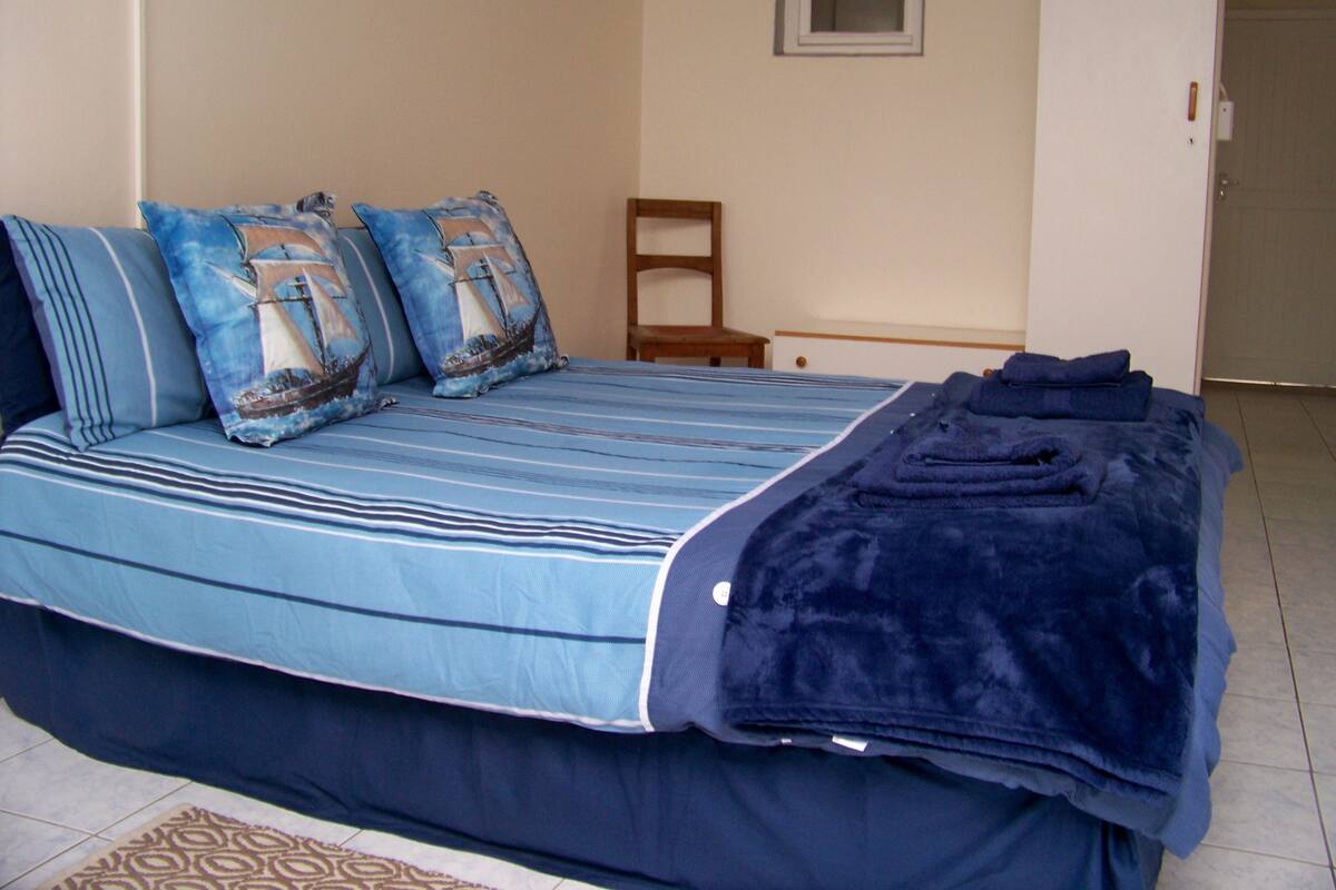 A bed with striped bedding and pillows, a dark blue blanket, a wooden chair, and a small window.