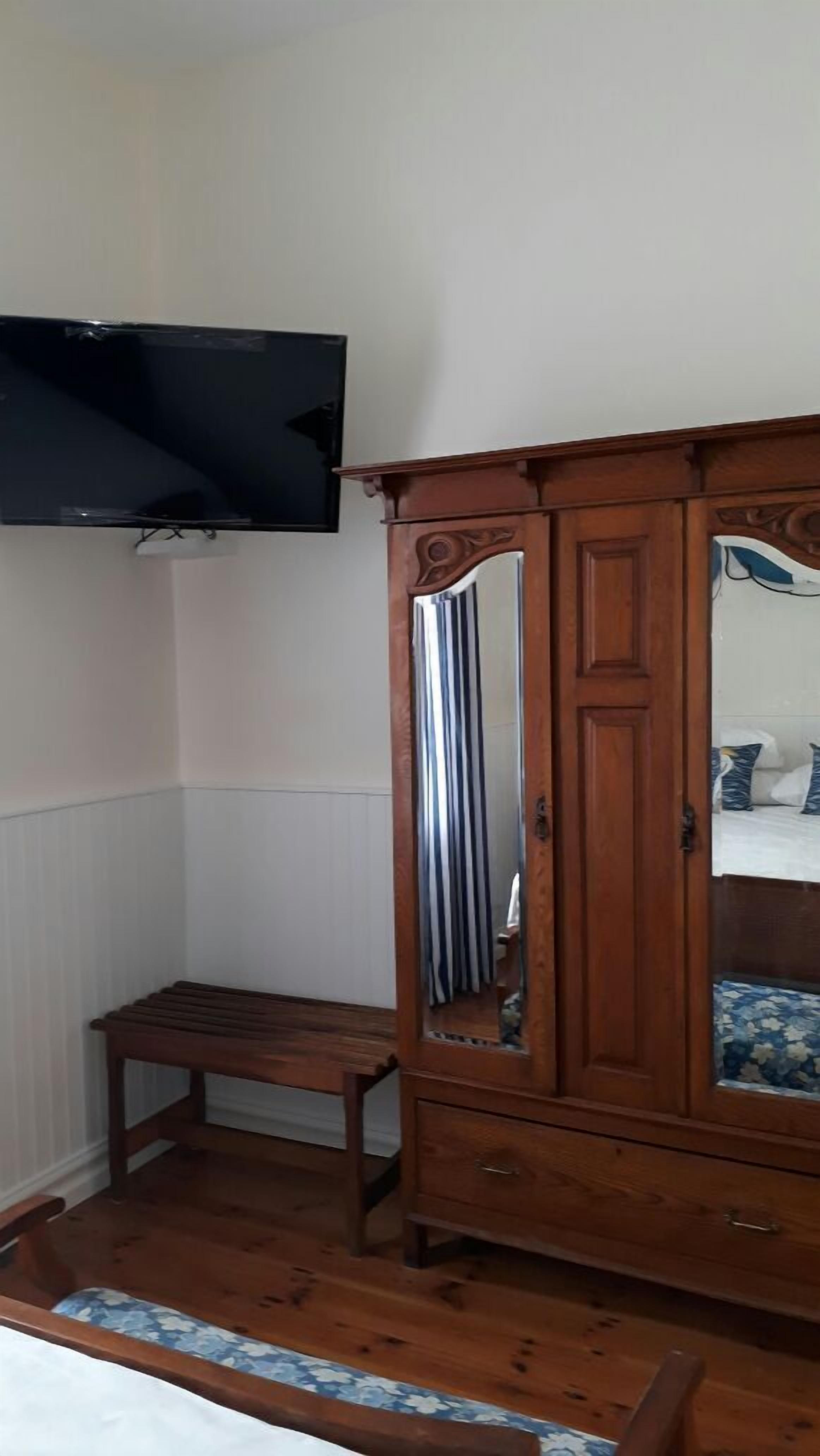 A bedroom with a wooden wardrobe, a bench, a television mounted on the wall, and a bed with a floral-patterned bedspread.