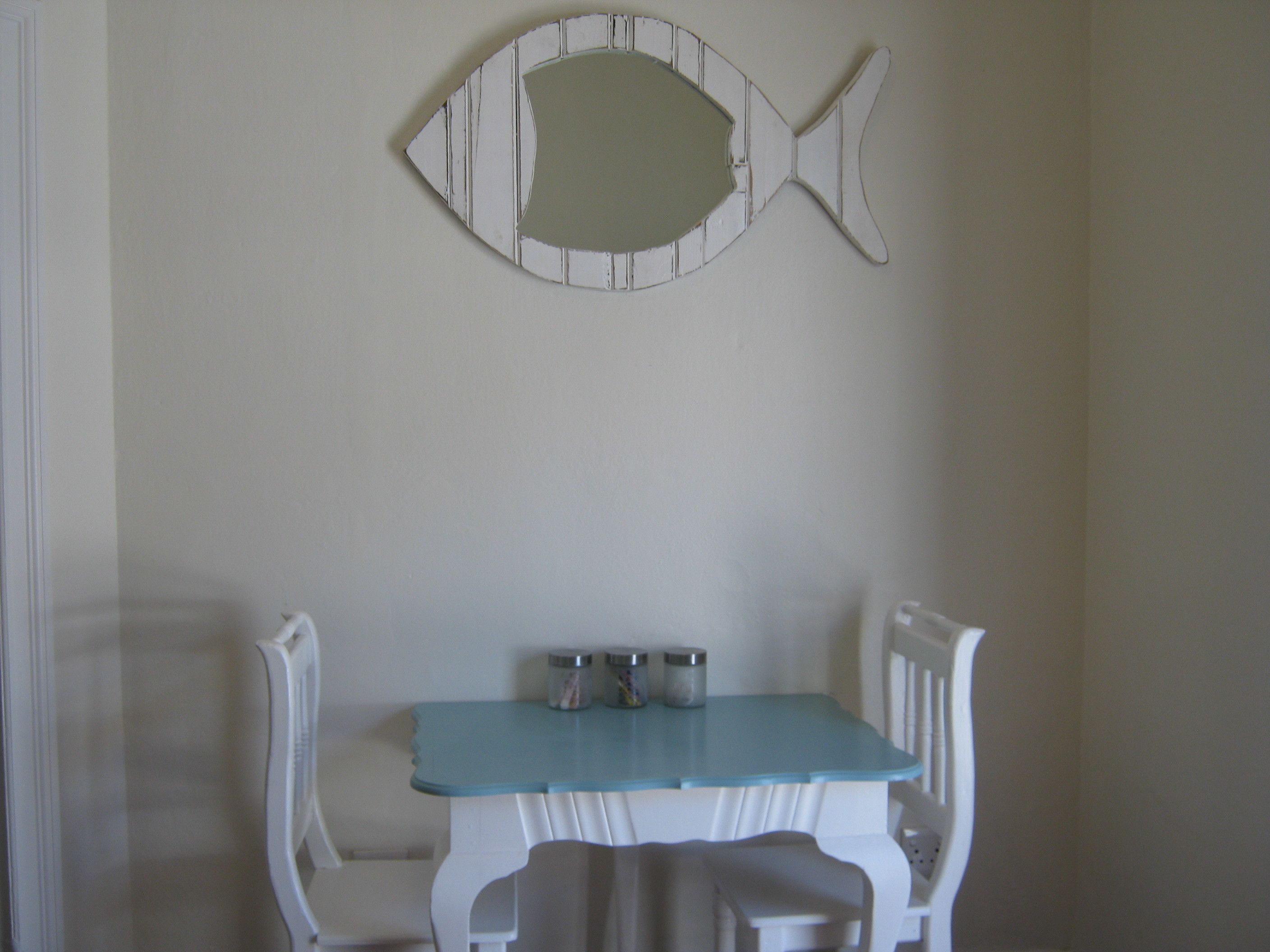 A dining area with a white table and chairs, a fish-shaped mirror on the wall, and two salt and pepper shakers on the table.