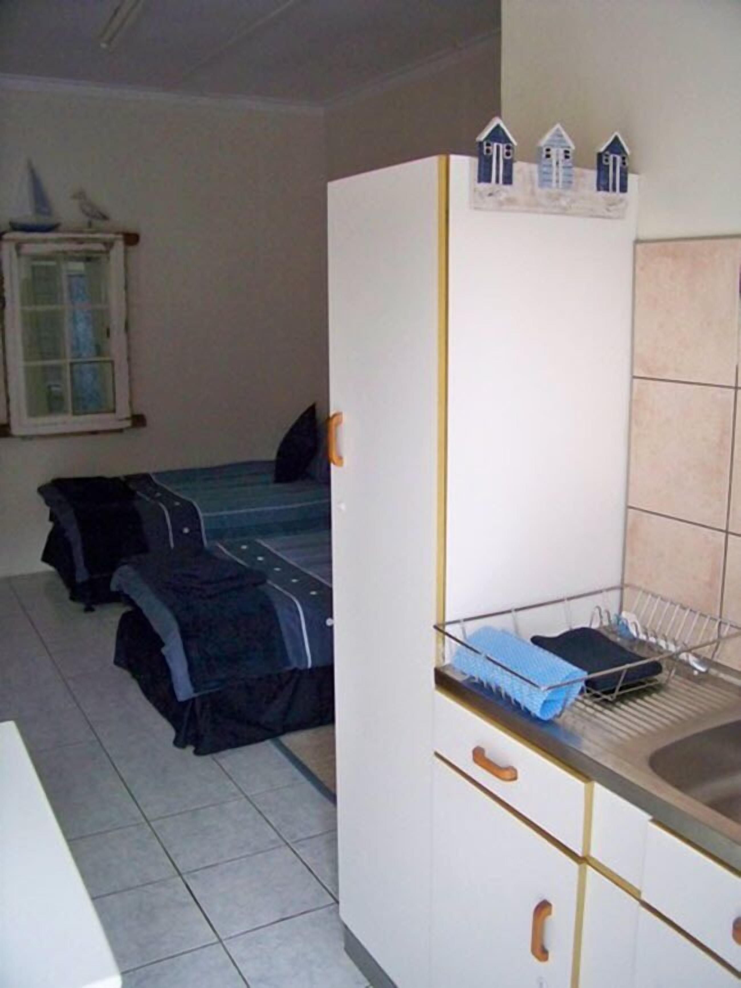 Una habitación pequeña y compacta con kitchenette, una cama con cubierta oscura y una ventana que da a un edificio.