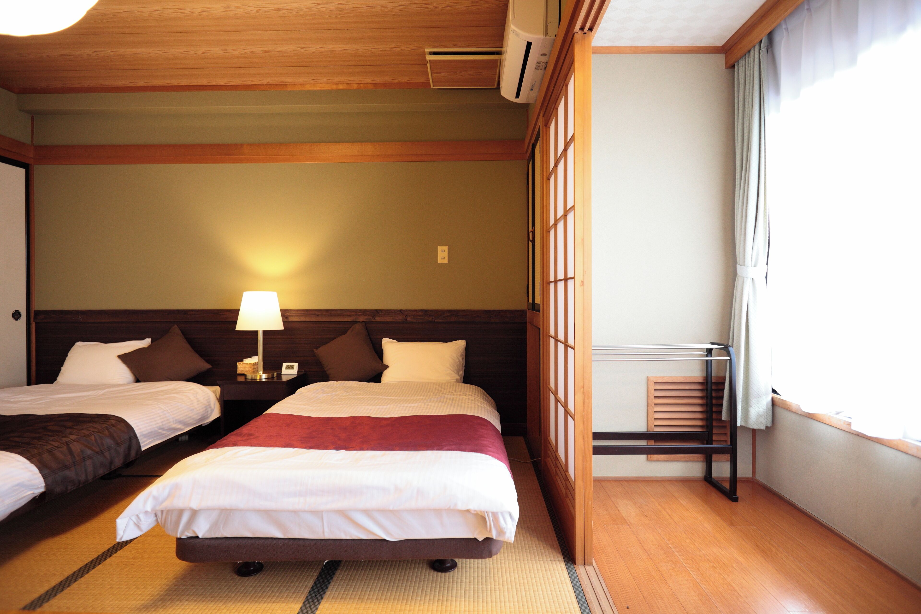 A hotel room with two beds, a nightstand with a lamp, a wooden ceiling, and a window with curtains.