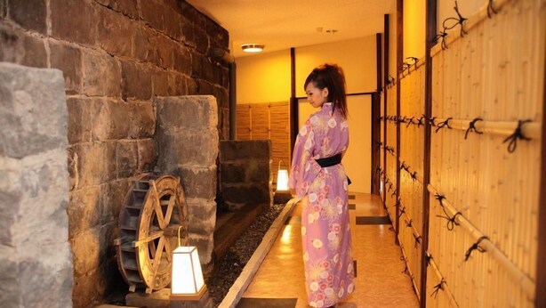 A traditional Japanese interior with a stone wall, a wooden water wheel, and a person in a kimono. - Yakushikan (Komoro)