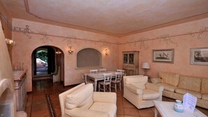 A spacious living room with a dining area, featuring a large arched window, a patterned sofa set, and a decorative wall with framed artwork. - Cala del Faro 103 (Arzachena)
