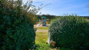 A playground with a slide and climbing structures, surrounded by dense greenery and a clear sky. - Cala del Faro 103 (Arzachena)