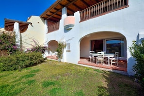 A white building with a patio area featuring a table and chairs, surrounded by greenery and a clear blue sky.