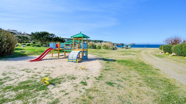 A playground with a slide and climbing structures, surrounded by a grassy area and a dirt path leading to a scenic view. - Cala del Faro 103 (Arzachena)