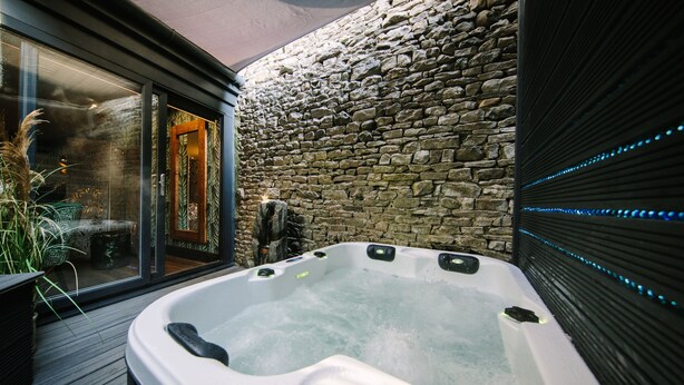 A hot tub with a stone wall backdrop and a wooden deck. - Absoluxe Suites (Carnforth)