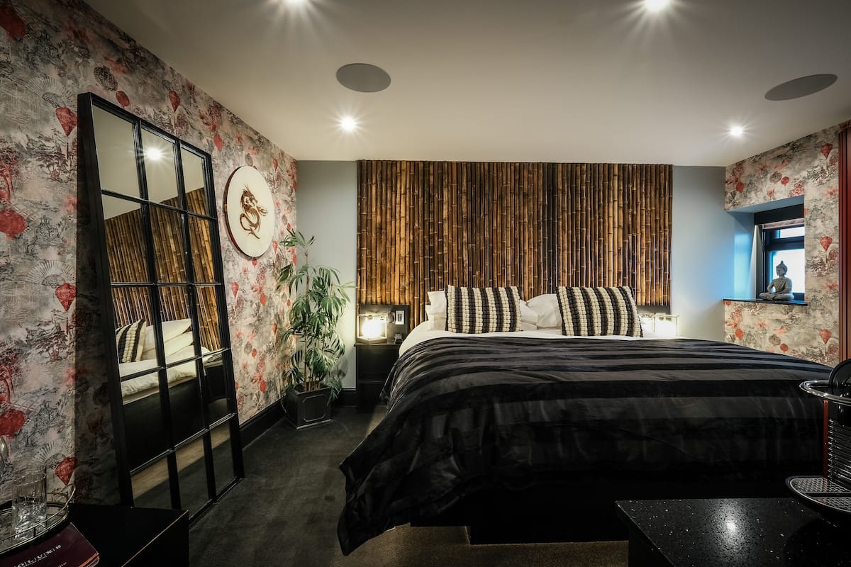 A modern bedroom with a large bed, a mirror, a wall-mounted TV, and a decorative plant.