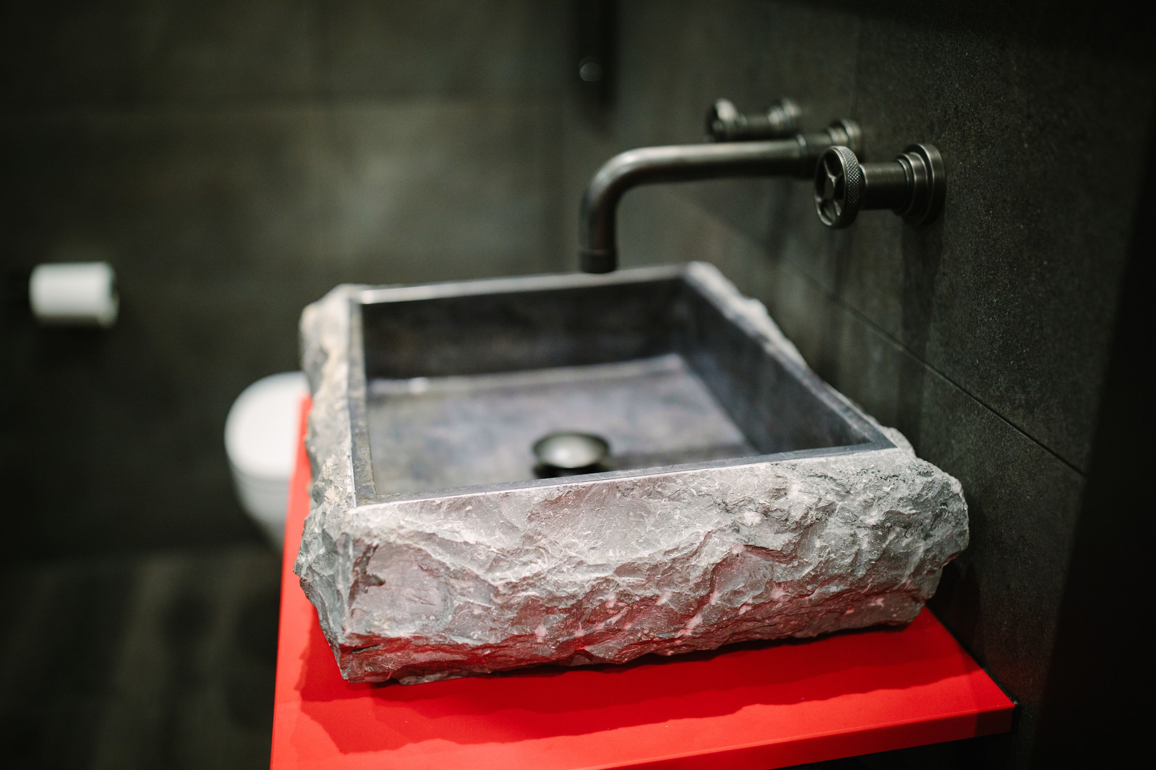 A bathroom with a stone sink on a red platform, a dark tiled wall, and a visible toilet in the background.