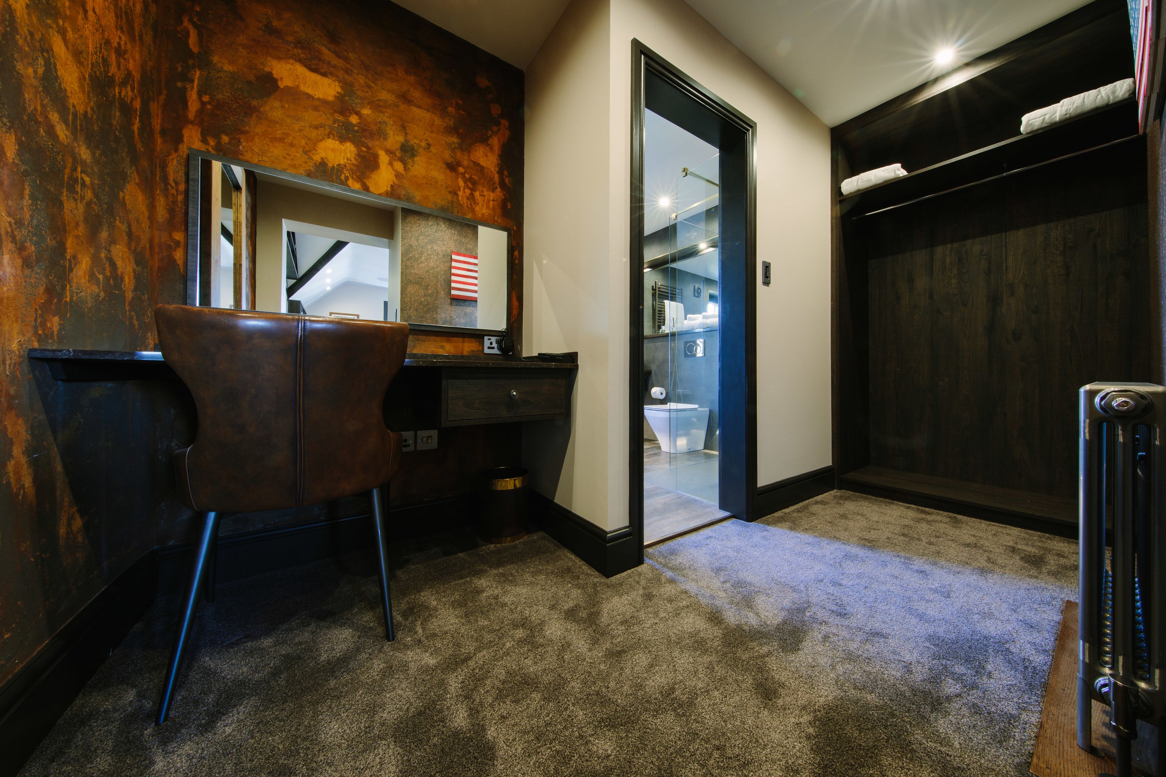 A room with a desk, chair, and a large mirror. There is a door leading to a bathroom with a toilet and a shower. The room has a carpeted floor and a dark wooden wardrobe.