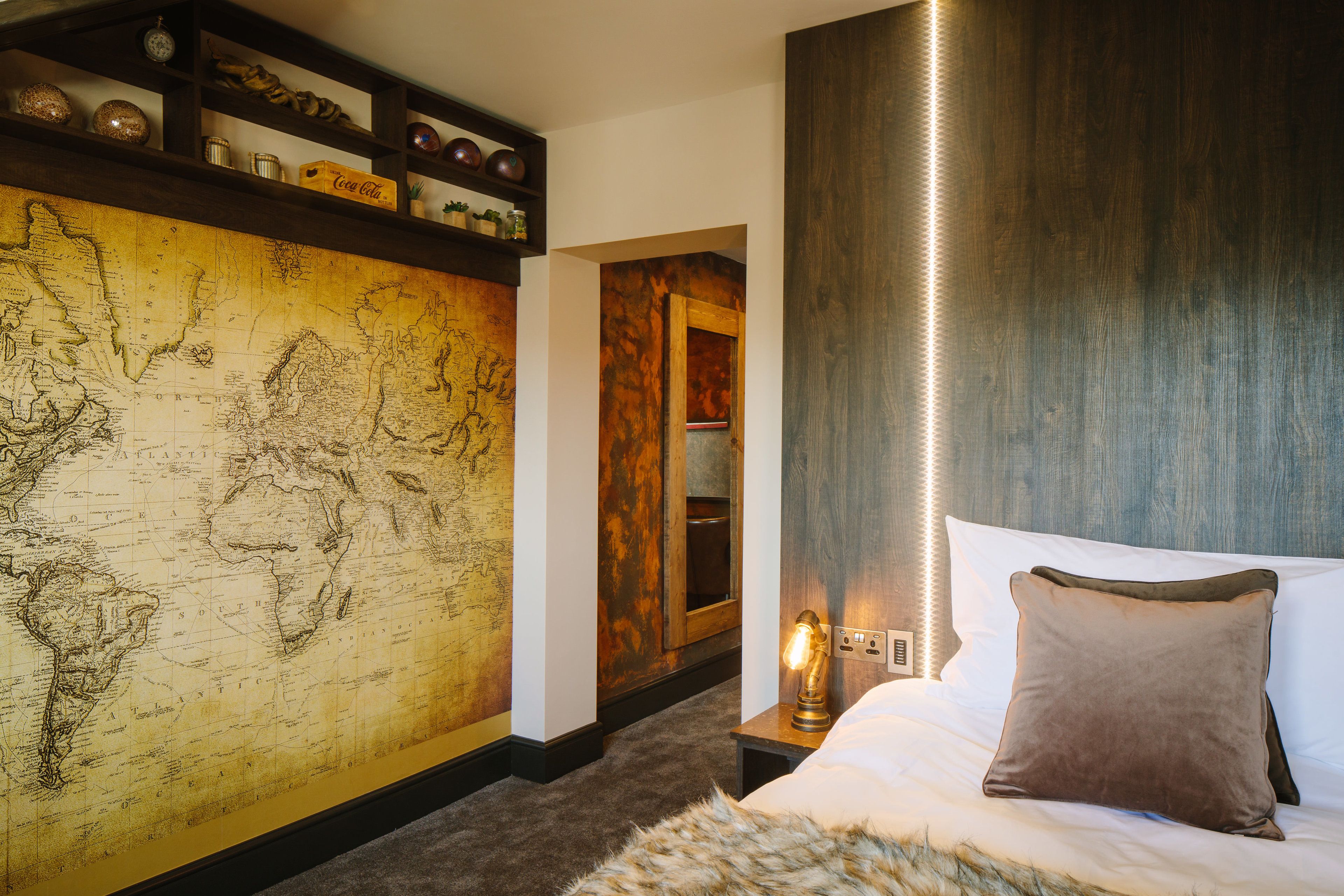 A hotel room with a large world map on the wall, a wooden door, a bedside lamp, and a bed with a brown pillow.