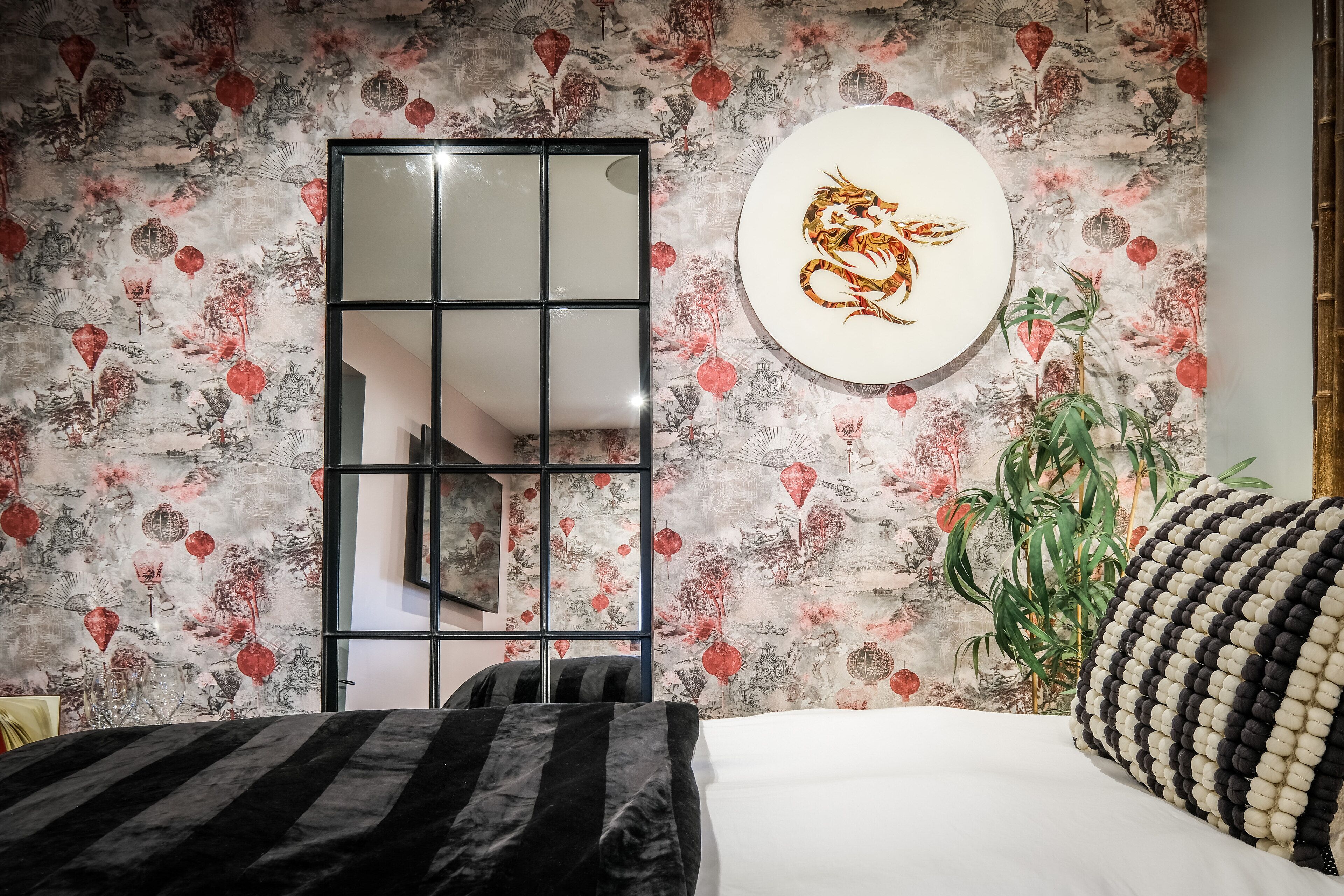 A bedroom with a bed, a large mirror, a wall with traditional artwork, and a potted plant.