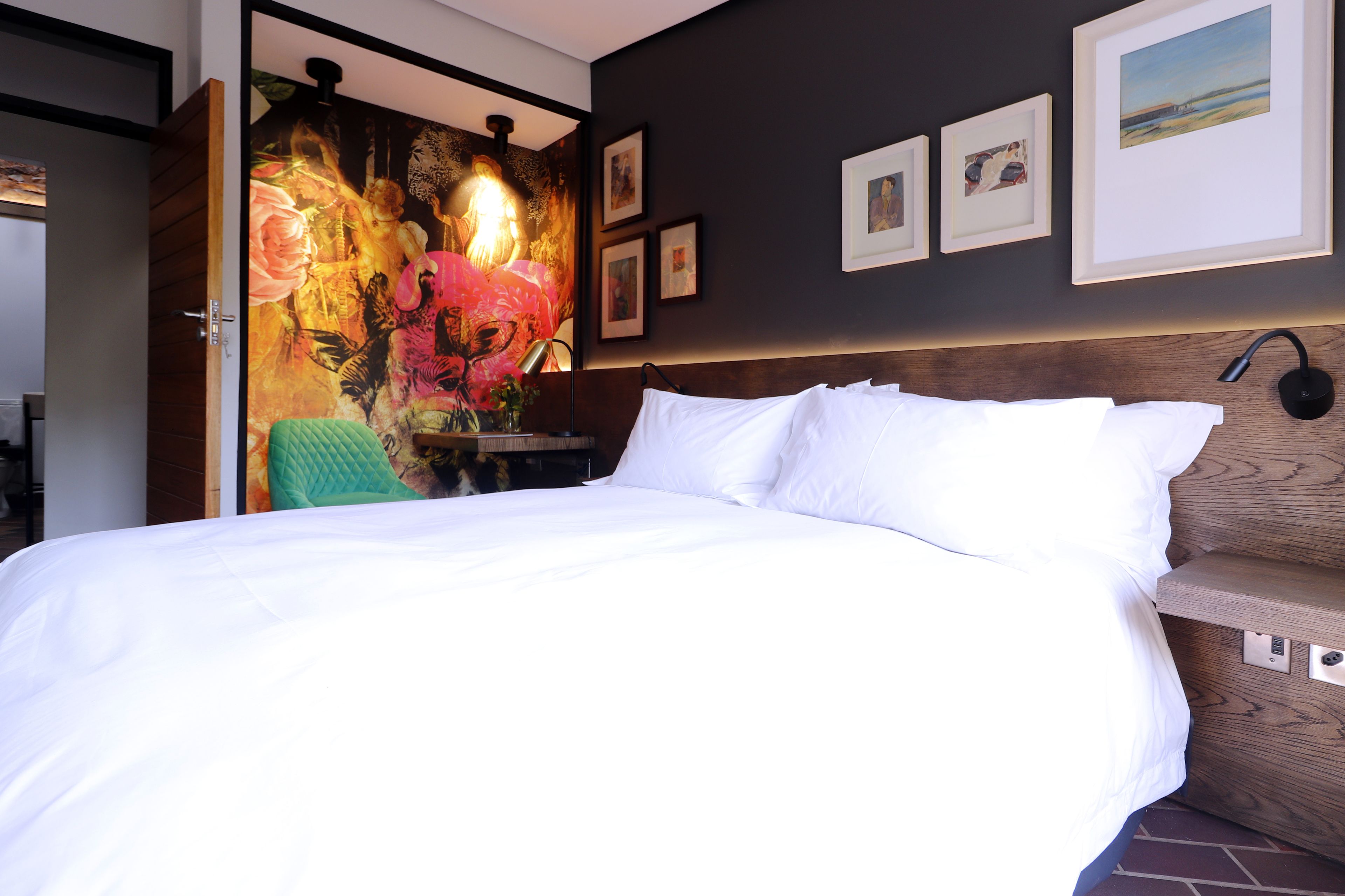 Une chambre d’hôtel avec un grand lit, une tête de lit en bois, un mur orné de tableaux encadrés et une fresque murale aux couleurs vives.