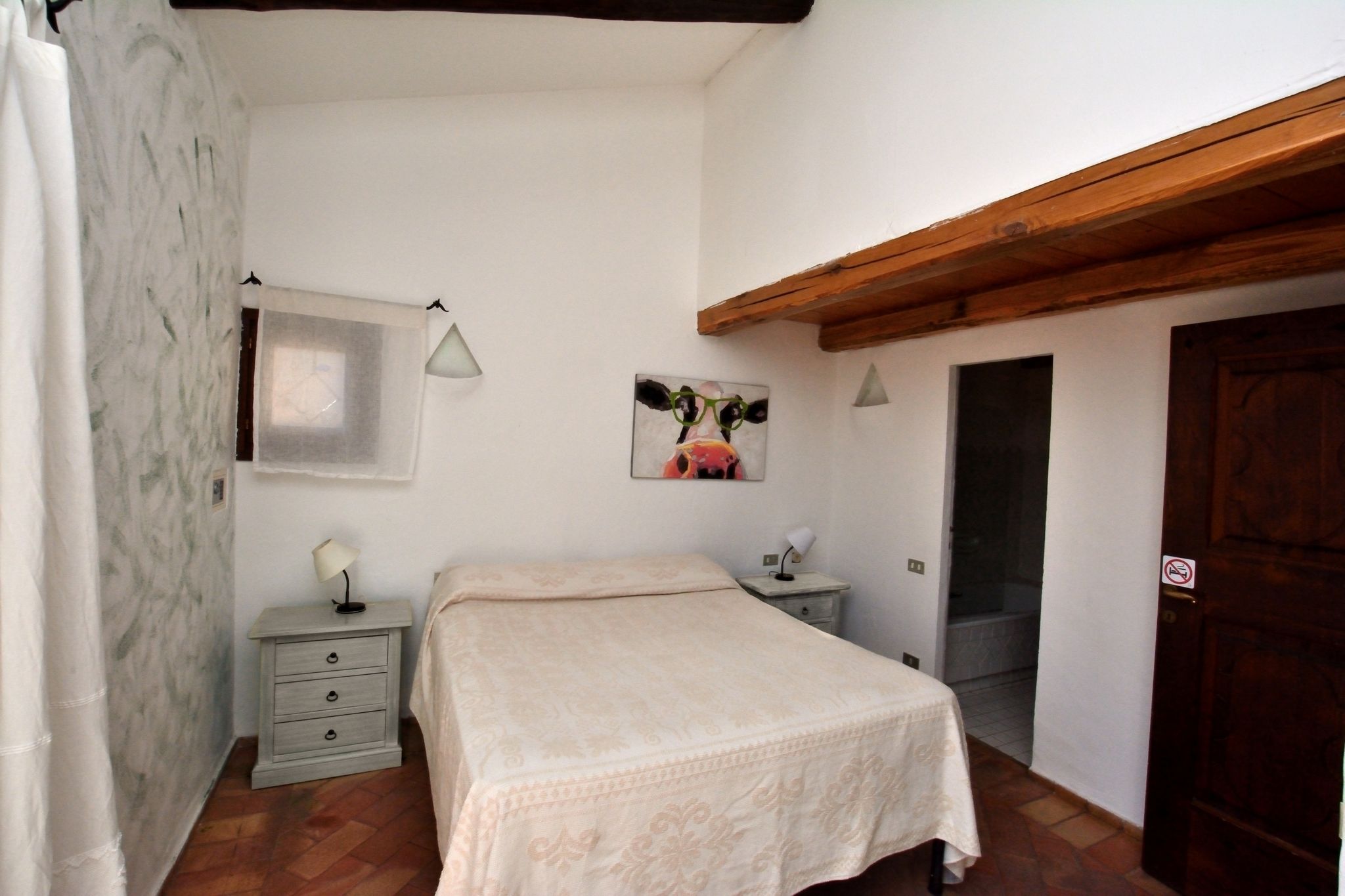 A bedroom with a bed, a nightstand, a lamp, a picture on the wall, and a wooden door.