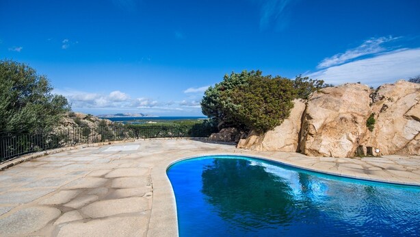 Una piscina de borde curvo, rodeada de baldosas de piedra y una cerca de metal.