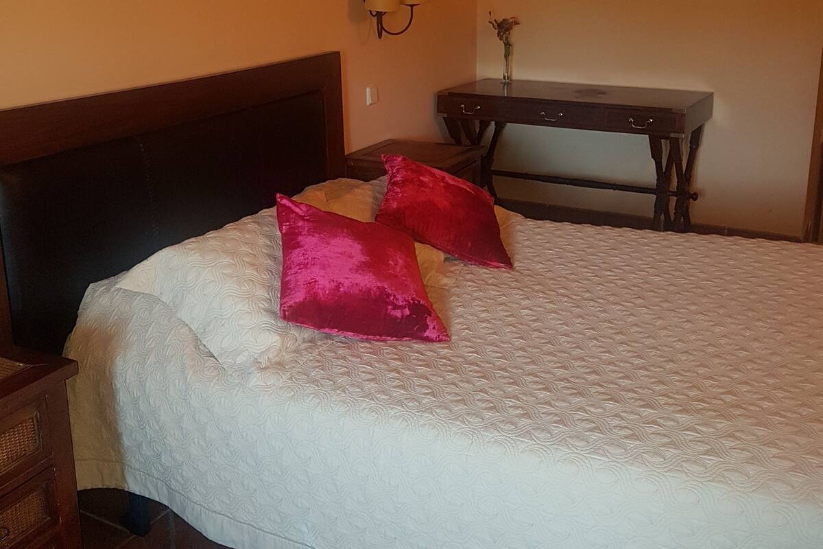A bedroom with a large bed, a nightstand, a dresser, a small table with a vase, and a wall-mounted lamp.
