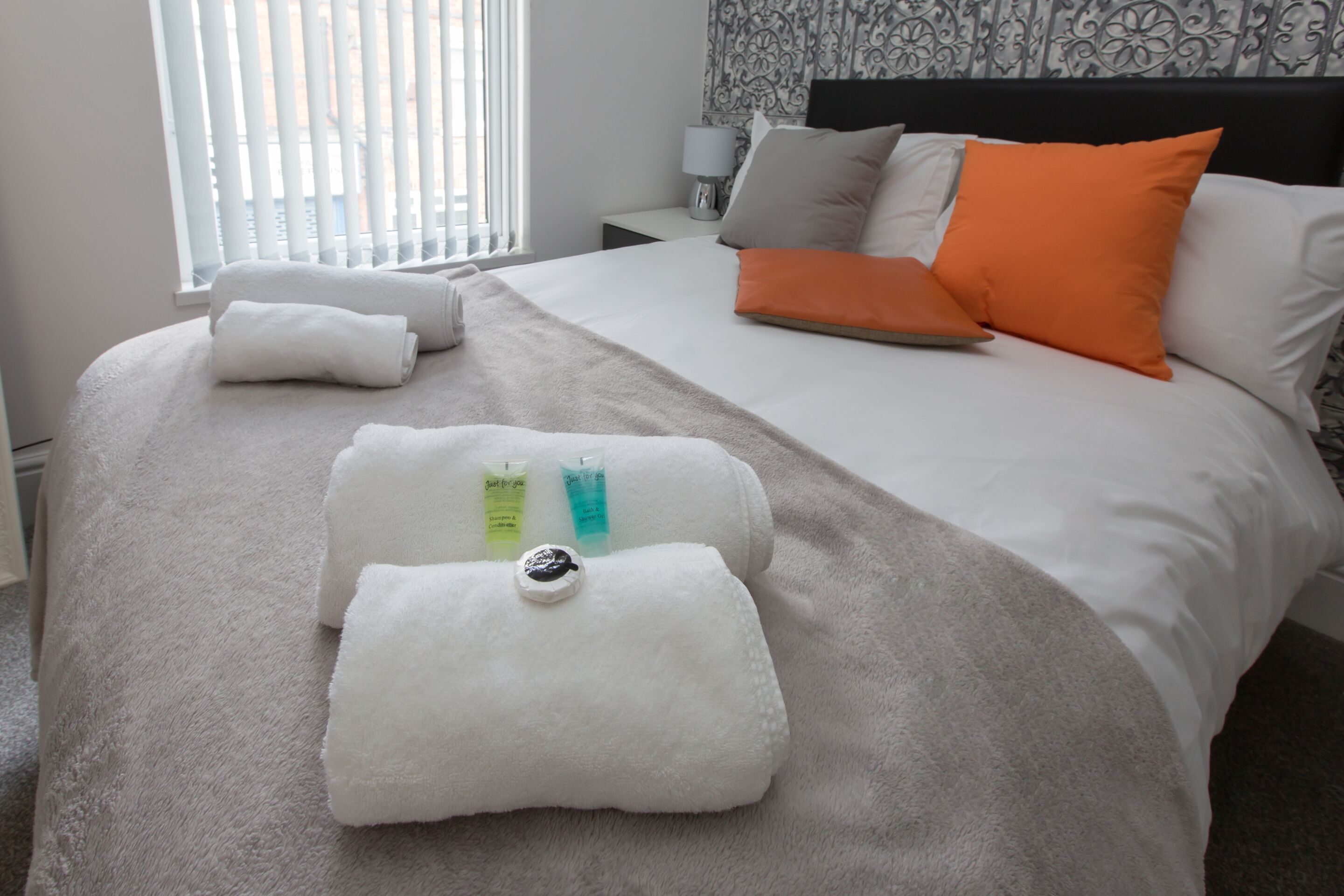 A neatly made bed with white and orange pillows, a grey blanket, and two rolled towels with toiletries on them.