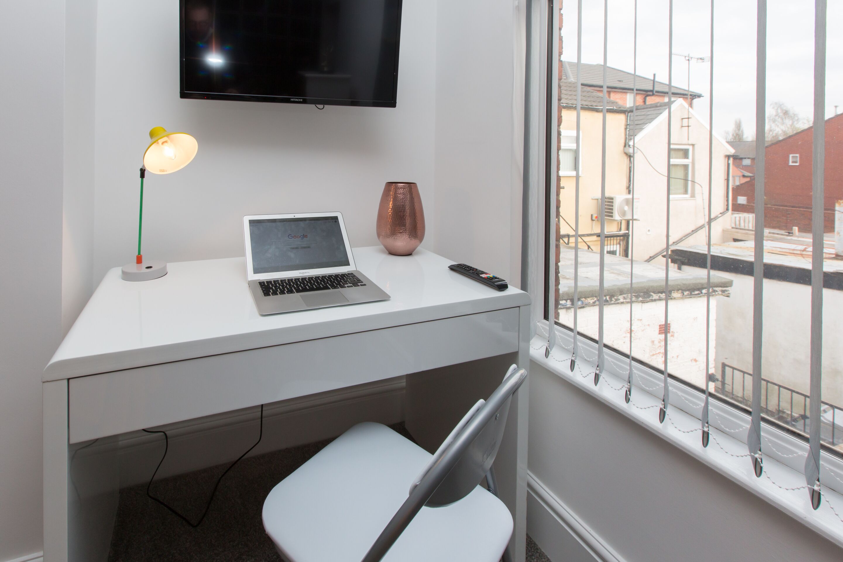 Un espace de travail moderne avec un ordinateur portable, une lampe et un vase posés sur un bureau blanc près d’une fenêtre ornée de barreaux métalliques.