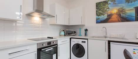 A modern kitchen with white cabinets, a microwave, a kettle, a washing machine, and a sink.
