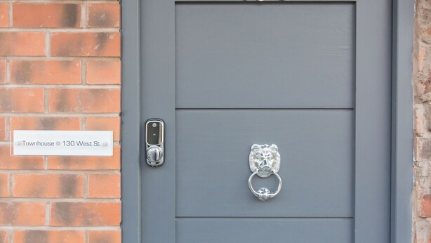Une porte grise portant le numéro 130, un heurtoir métallique et une plaque indiquant « Maison de ville, 130, rue West ».