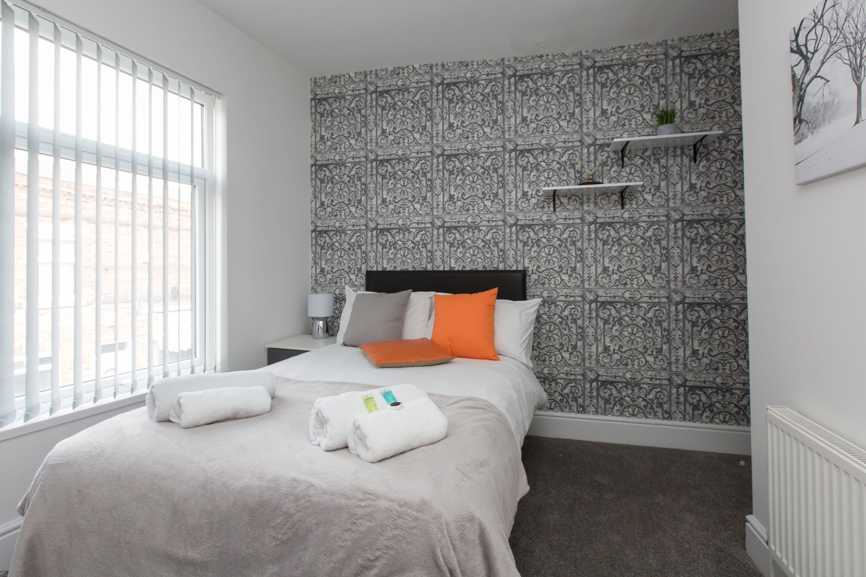 A hotel room with a bed, a window with blinds, a patterned wallpaper, and a small shelf with a plant.