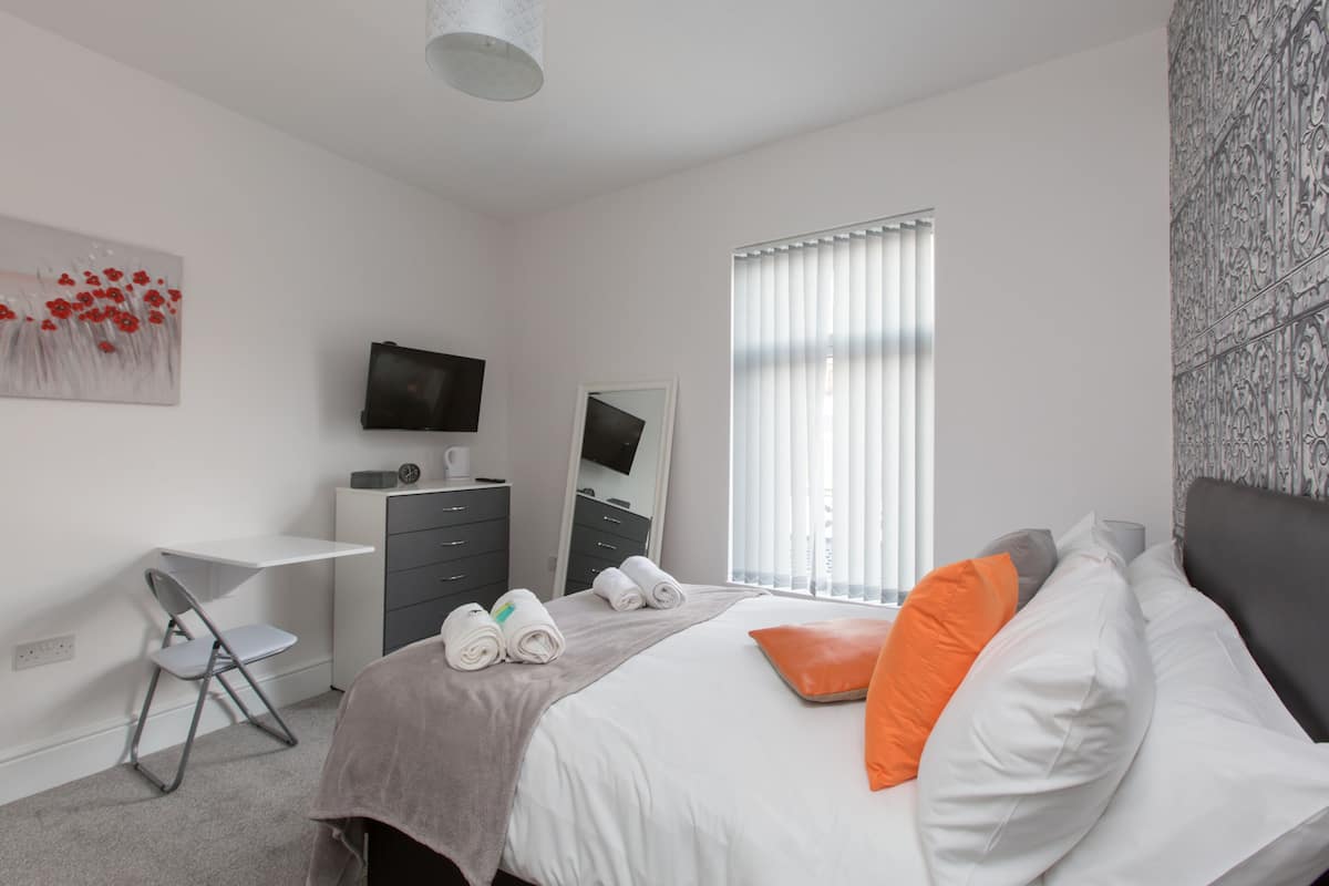 Een moderne slaapkamer met een bed, een bureau, een stoel, een televisie en een raam met jaloezieën.