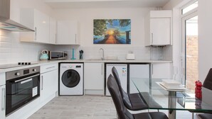 Une cuisine moderne avec des meubles blancs, un lave-linge, un four à micro-ondes et une table à manger en verre, accompagnée de chaises.