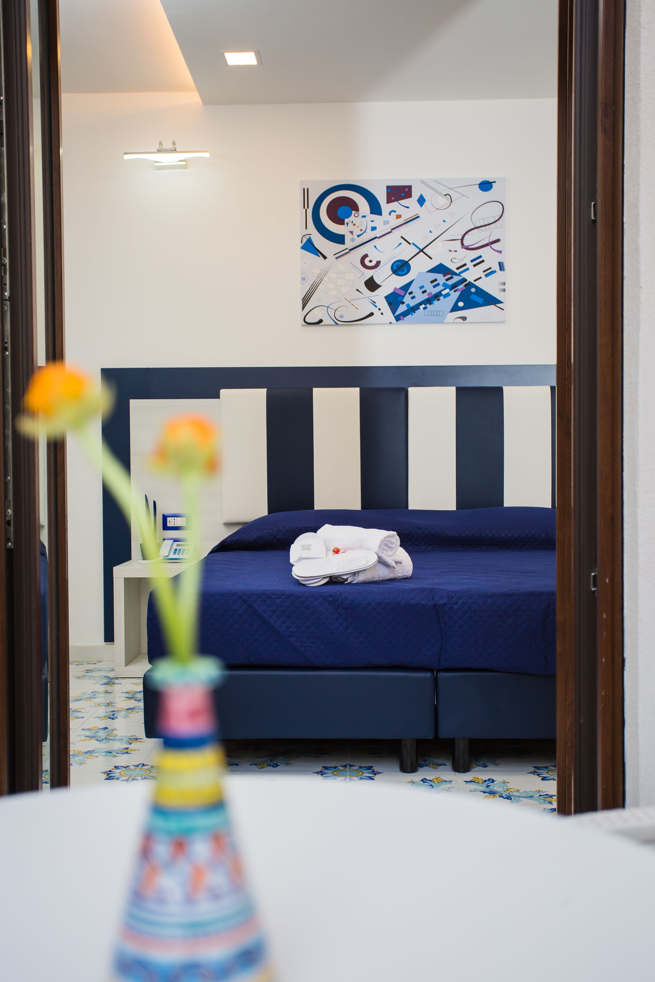 A hotel room with a bed, a bedside table, a wall art piece, and a vase with flowers.